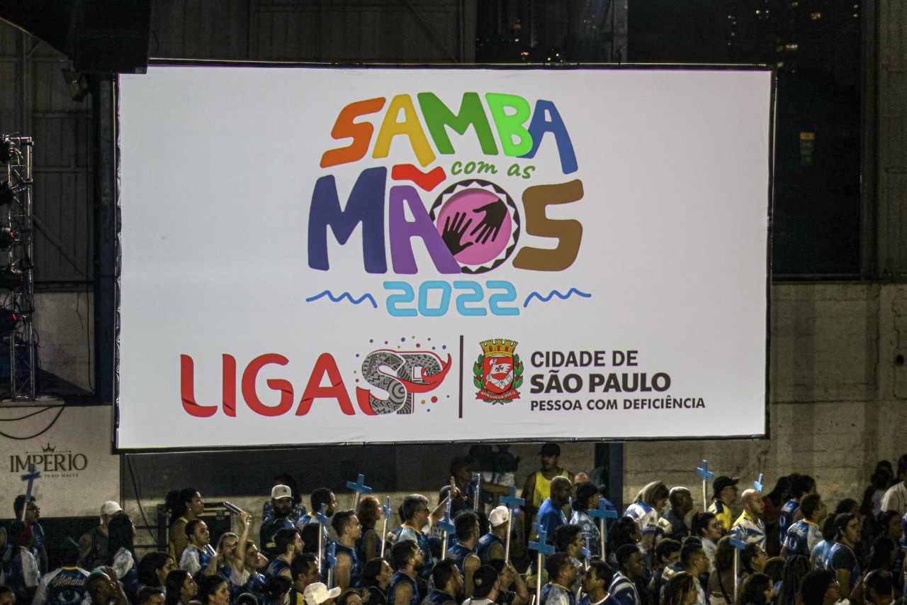 No telão, no centro a logomarca do projeto "Samba com as Mãos 2022". Abaixo o logotipo da Liga-SP e ao lado o logotipo Cidade de São Paulo Pessoa com Deficiência. Esse telão está instalado ao lado do palco na quadra da Águia de Ouro, abaixo dele é possível ver integrantes da escola.